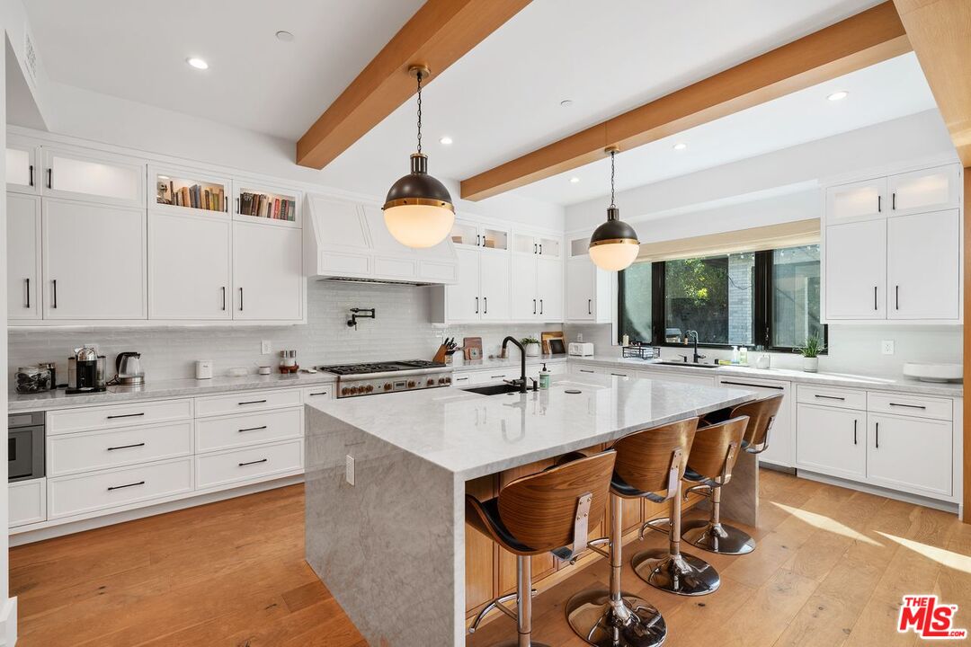 Kitchen with a large marble island, white cabinetry, wood ceiling beams, and three bar stools.