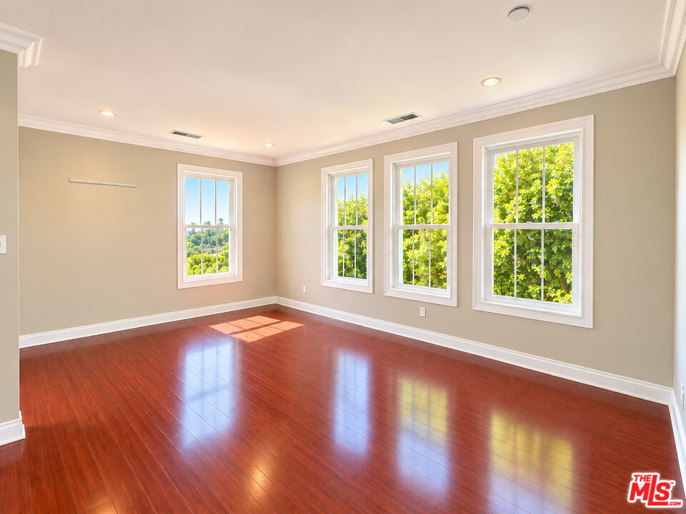 Vacant room with polished reddish hardwood floors and four windows overlooking greenery.