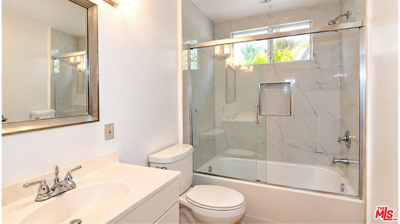 Bathroom with a white vanity, toilet, and glass-enclosed tub against marble-patterned tile.