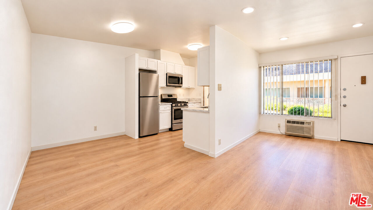Open-plan apartment living area with light wood floors, a compact stainless-steel kitchen, and a window with vertical blinds.