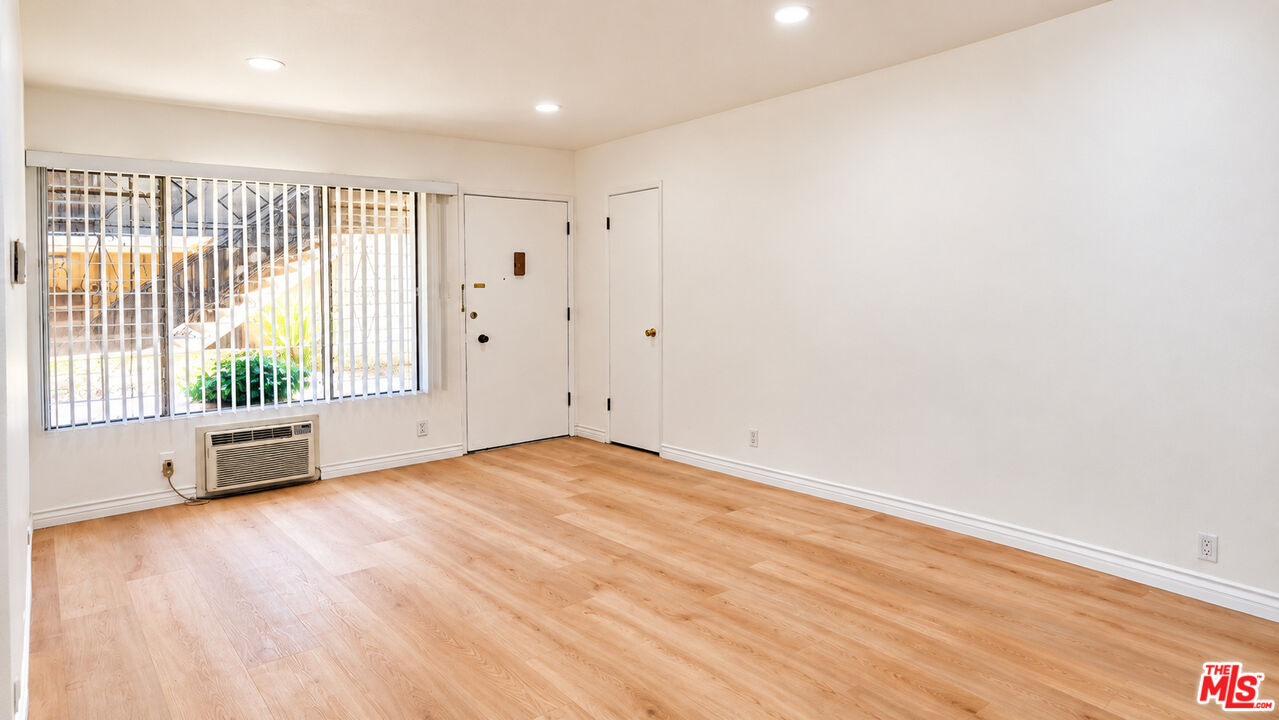 Vacant living room with light wood floors, a barred window, and a wall-mounted air conditioner.