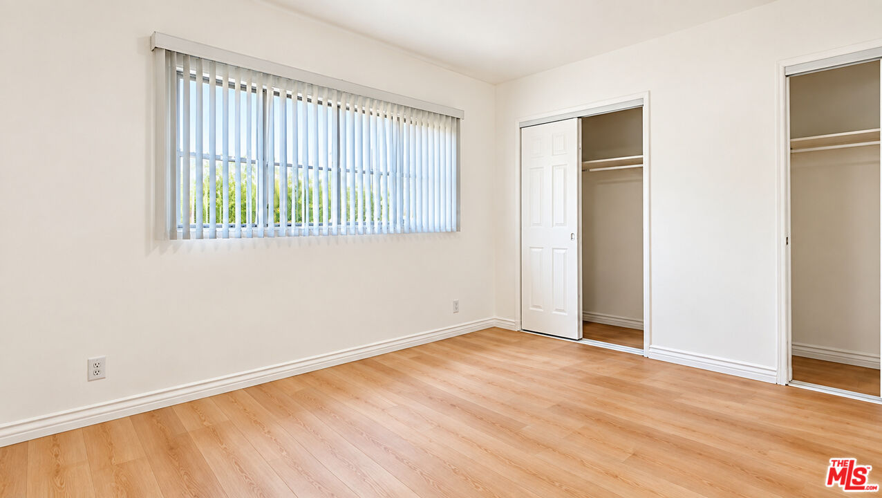 Empty bedroom with light wood floors, vertical blinds, and two open closets.