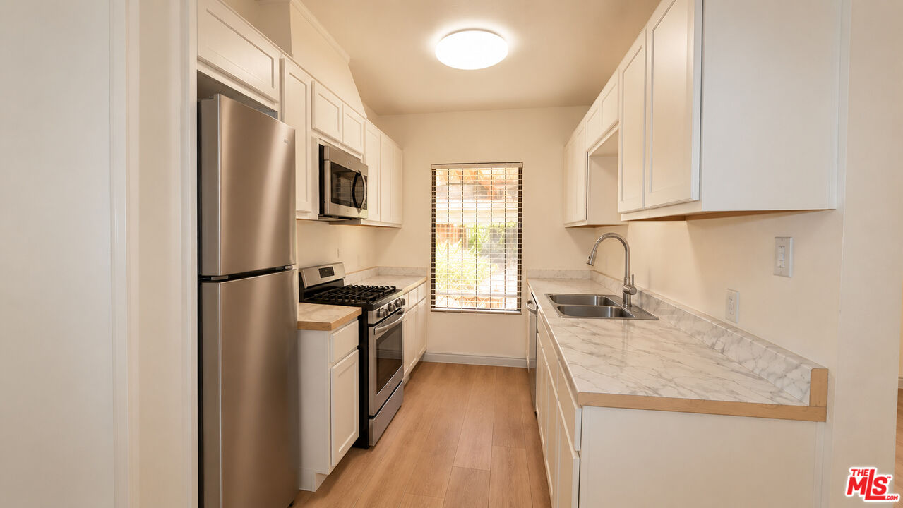 Galley kitchen with white cabinets, stainless steel appliances, marble-look counters, and a window with blinds.