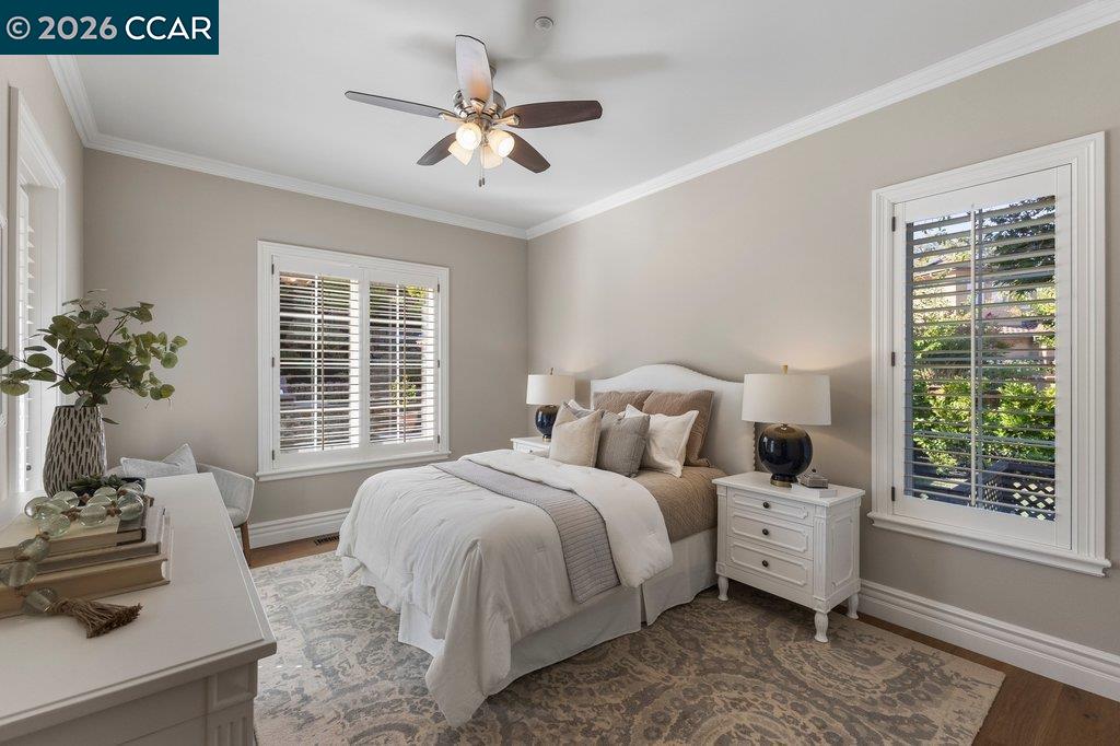 Staged bedroom with a white bed, ceiling fan, and plantation shutters on two windows.