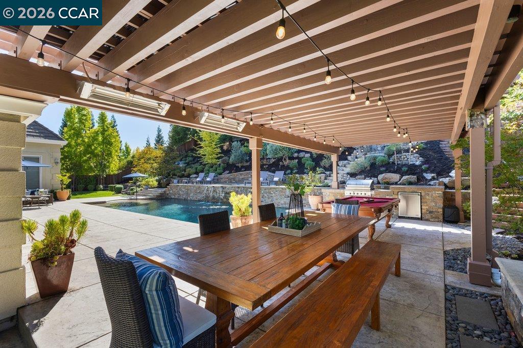 Covered patio with a long wooden dining table overlooking a swimming pool and landscaped yard.