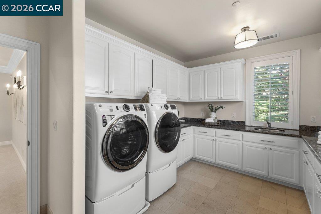 Laundry room with front-loading washer and dryer, white cabinetry, and a sink beneath a shuttered window.