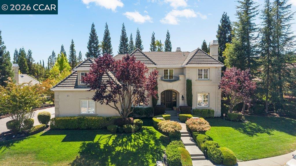 Two-story beige home with a tiled roof, arched entry, landscaped lawn, and mature trees.