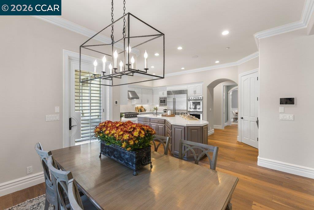 Dining area with a large wood table beneath a geometric chandelier, opening onto a kitchen.