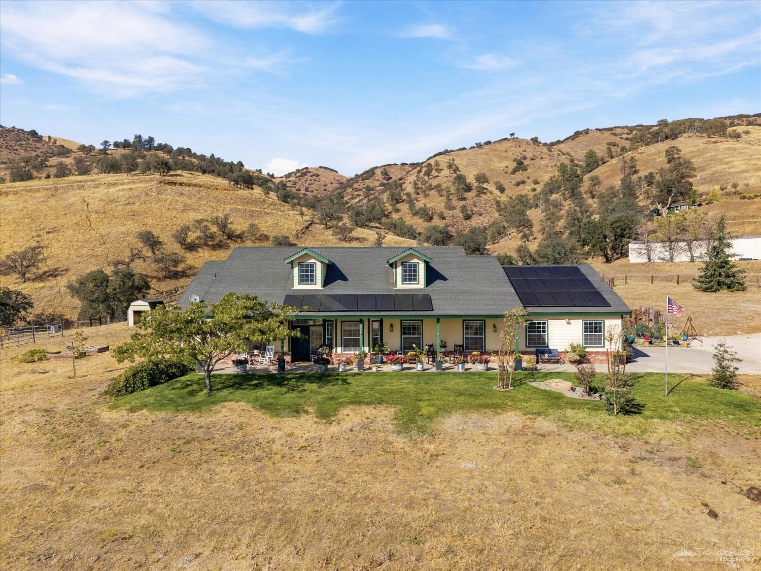 Ranch-style home with rooftop solar panels, a covered porch, and dry rolling hills behind it.