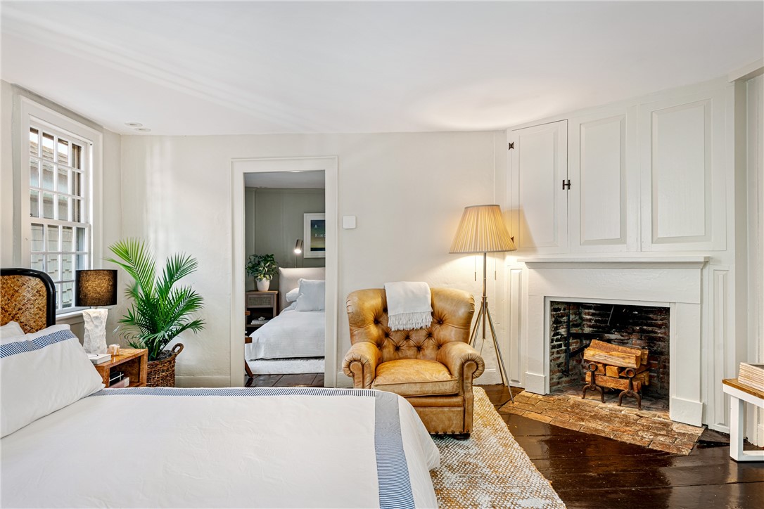 Bedroom with a white bed, leather armchair, and paneled fireplace.