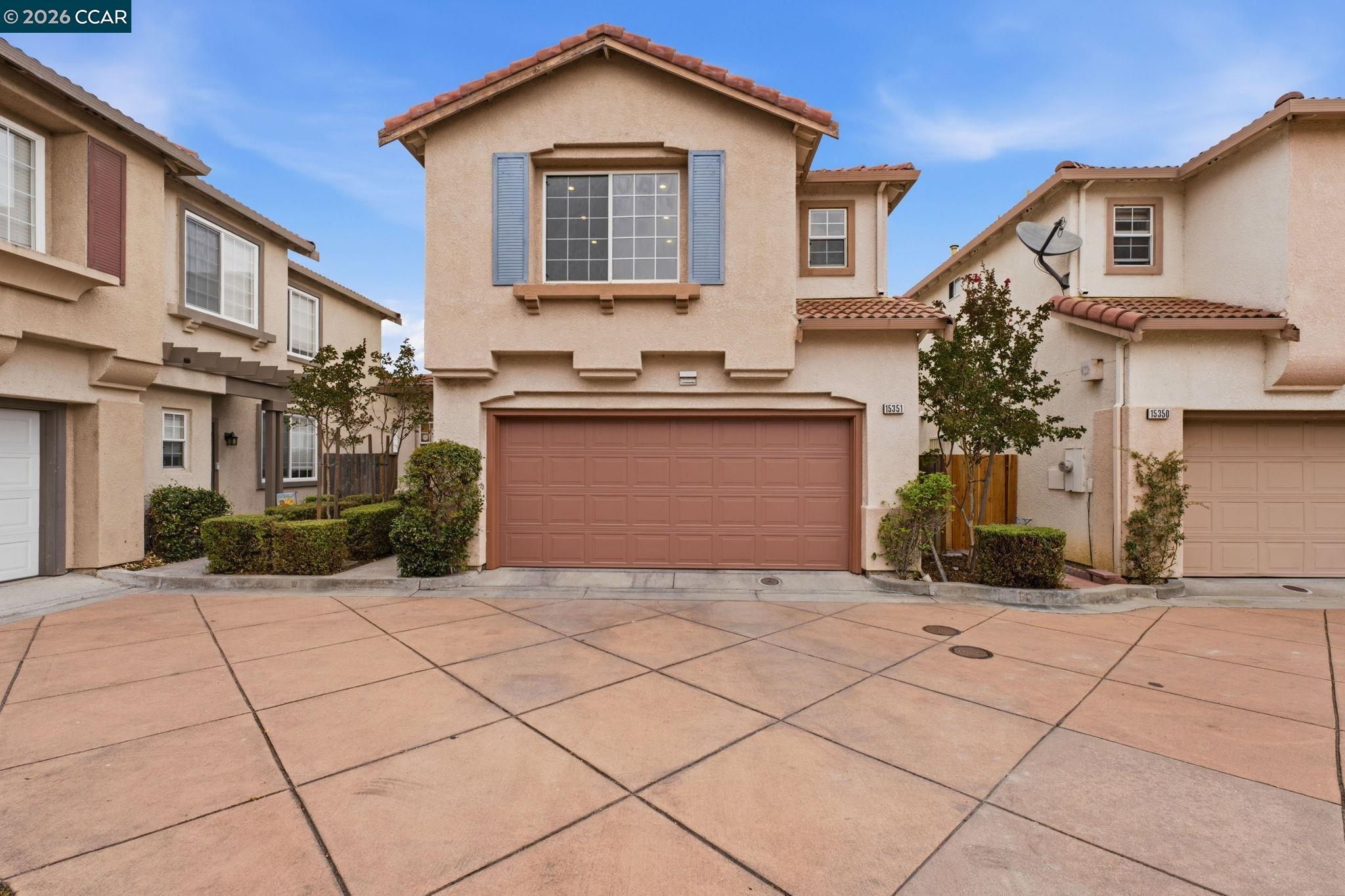 Two-story beige stucco home with blue shutters, a reddish garage door, and a broad paved driveway.