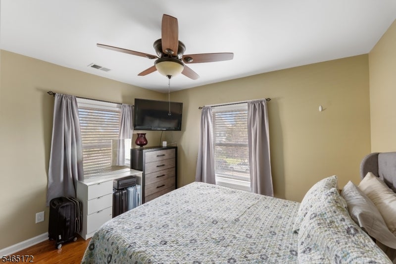 Furnished bedroom with patterned bedding, two curtained windows, and a ceiling fan.