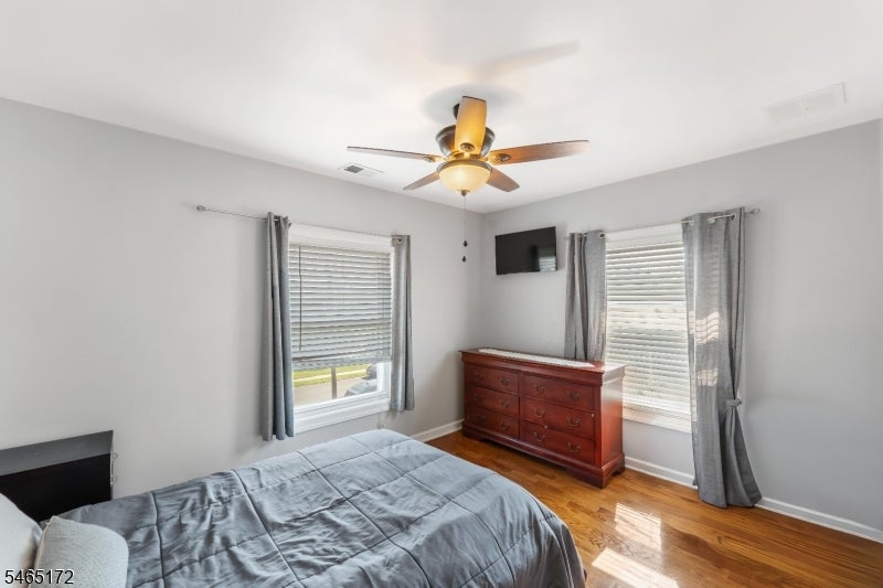 Bedroom with a gray bedspread, hardwood floors, and a wooden dresser between two curtained windows.