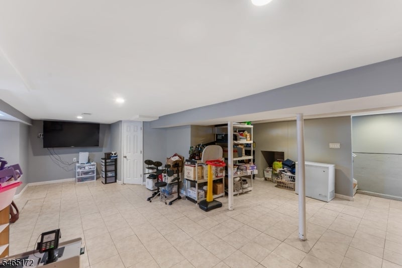 Finished basement with beige tile floors, exercise equipment, storage shelves, and a wall-mounted television.