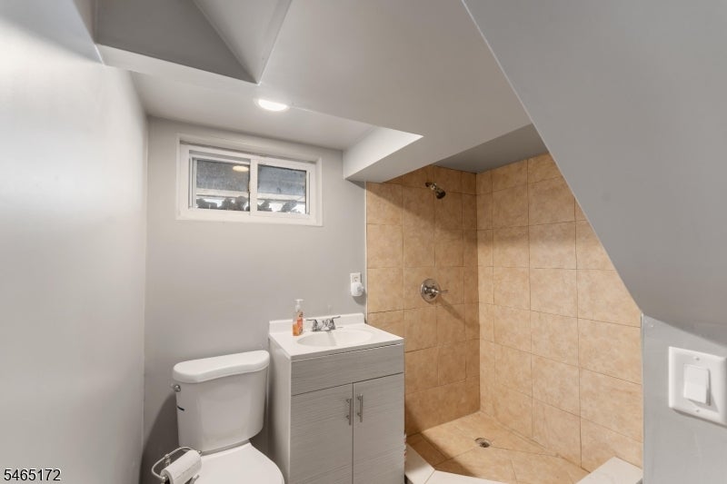 Bathroom with tan tiled shower, gray vanity, toilet, and small window beneath a sloped ceiling.