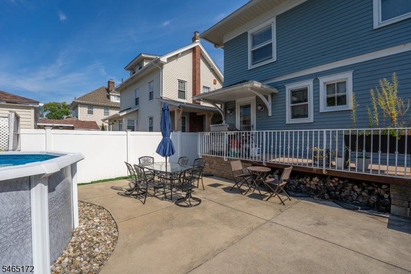 Backyard patio with outdoor dining furniture, raised deck, white fence, and above-ground pool beside a blue house.