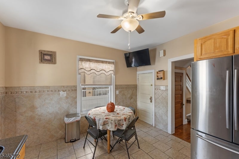 Kitchen dining area with a round floral-covered table, tiled walls, ceiling fan, and stainless-steel refrigerator.