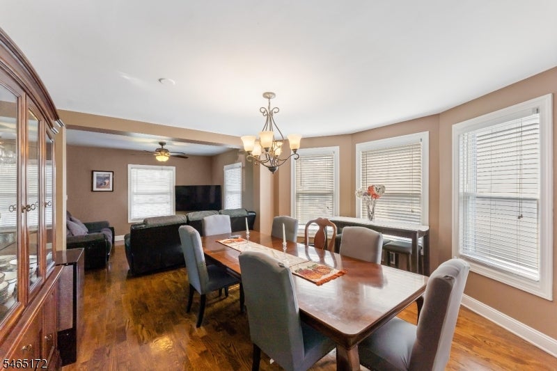 Dining room with a long wooden table, upholstered chairs, hardwood floors, and chandelier beside large windows.