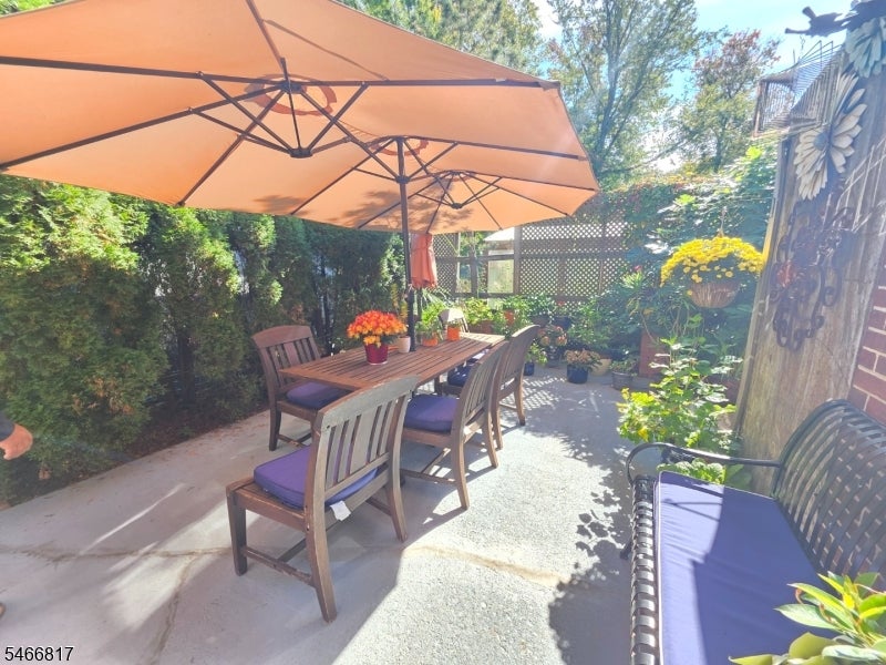 Patio with a wooden dining set beneath a large orange umbrella, surrounded by shrubs and potted flowers.