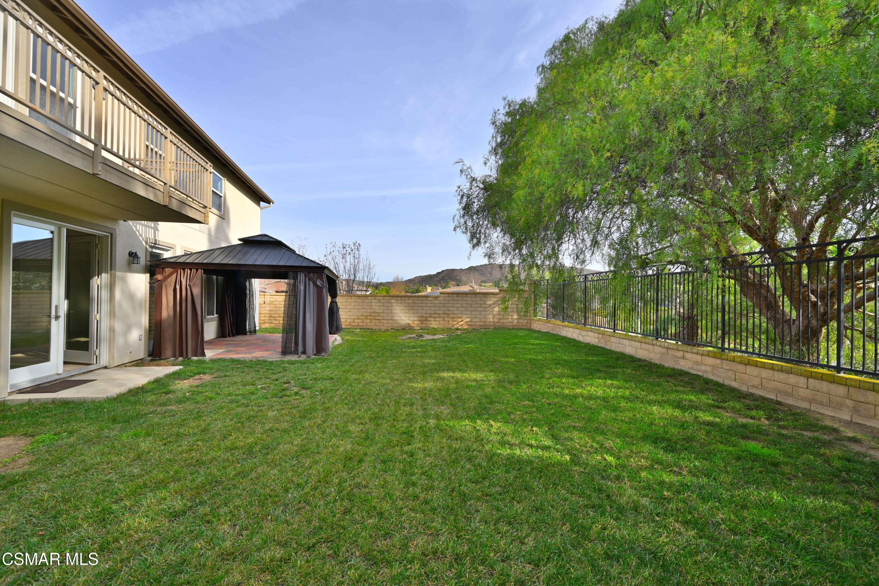 Backyard lawn with a curtained gazebo, mature tree, and two-story home with balcony.