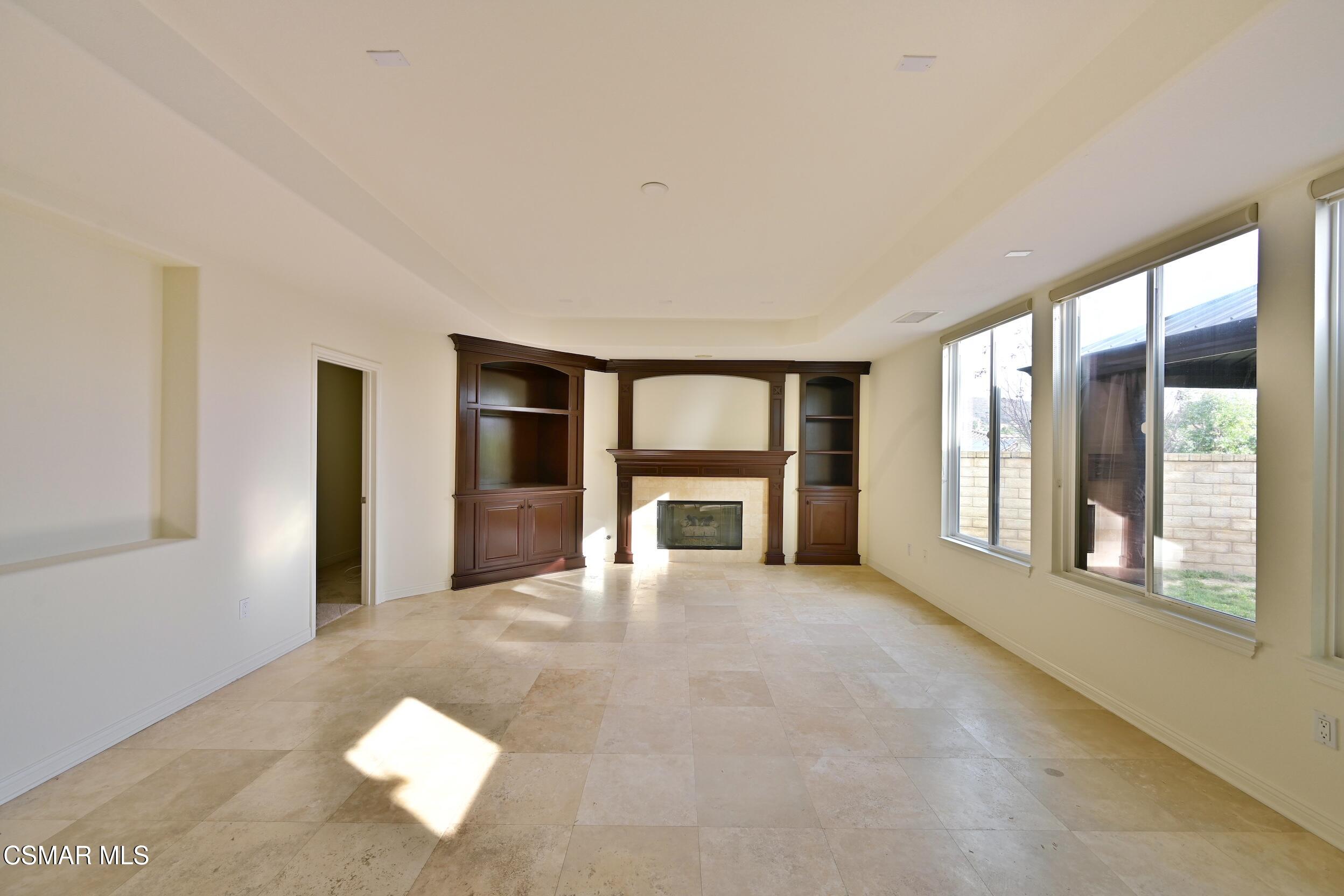 Empty living room with a dark wood fireplace and built-in shelves, pale tile floors, and large windows.
