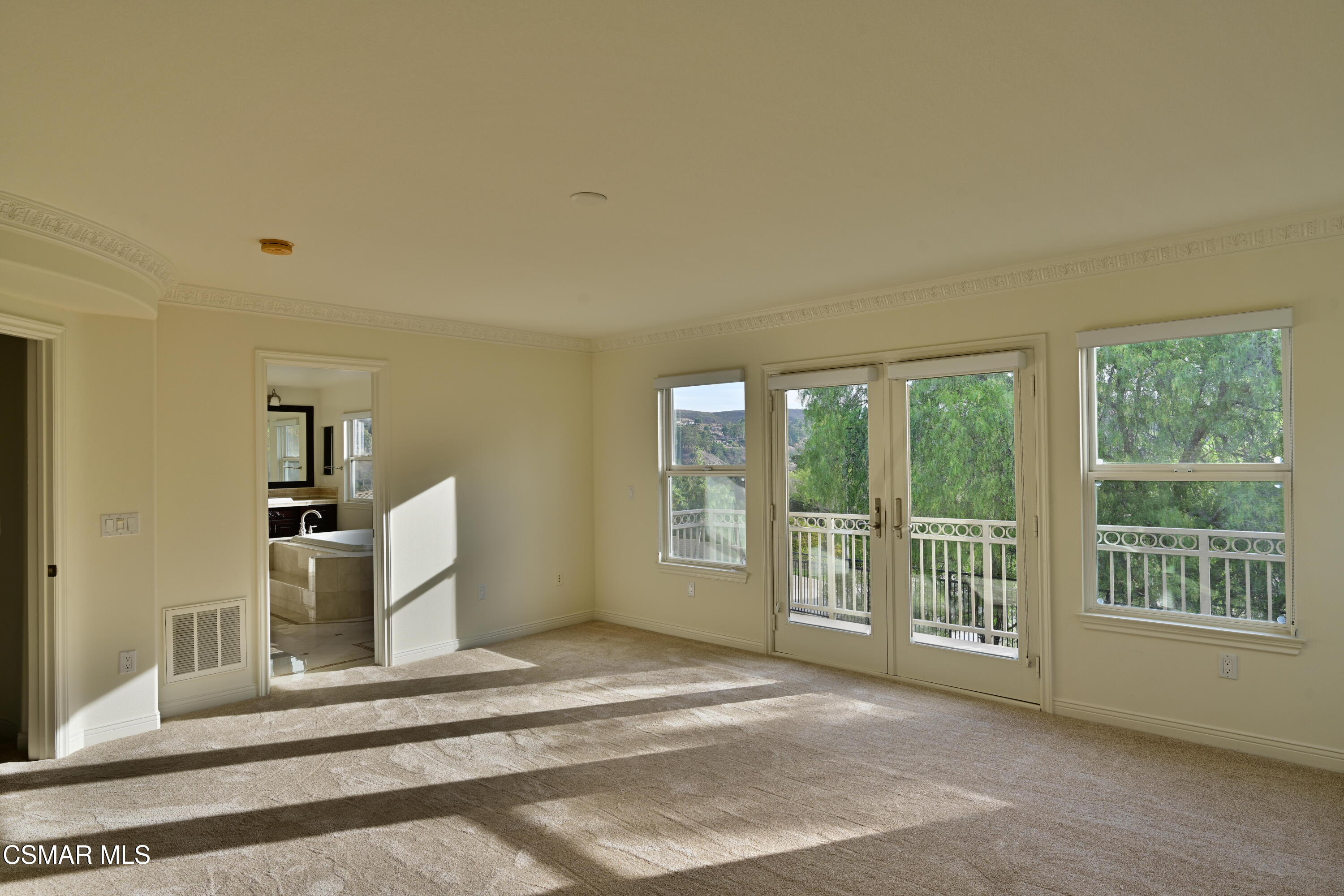 Empty bedroom with ornate crown molding, balcony doors, and sunlight across the carpet.