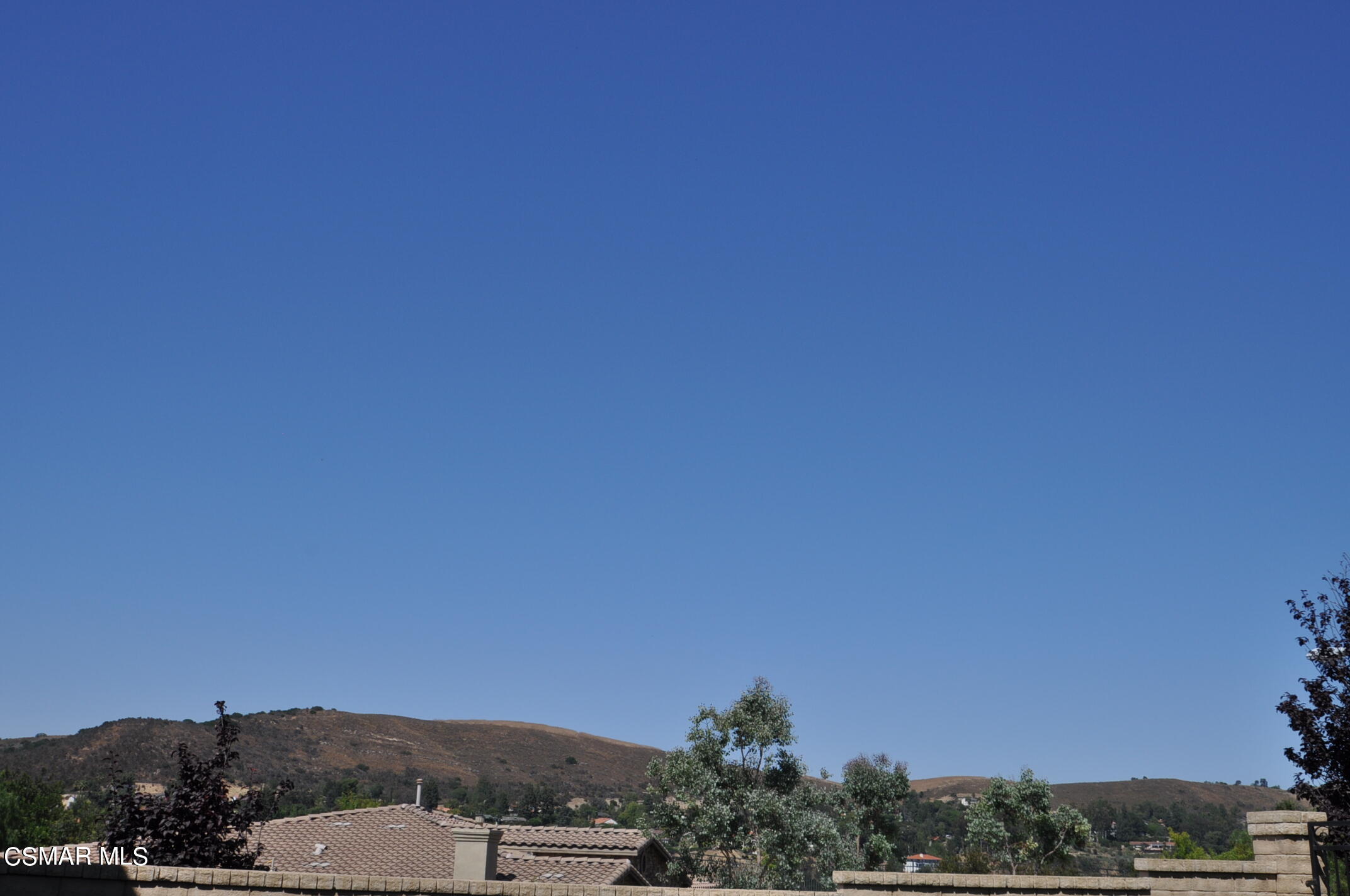 Rolling hills and trees beyond neighboring rooftops under a broad blue sky.