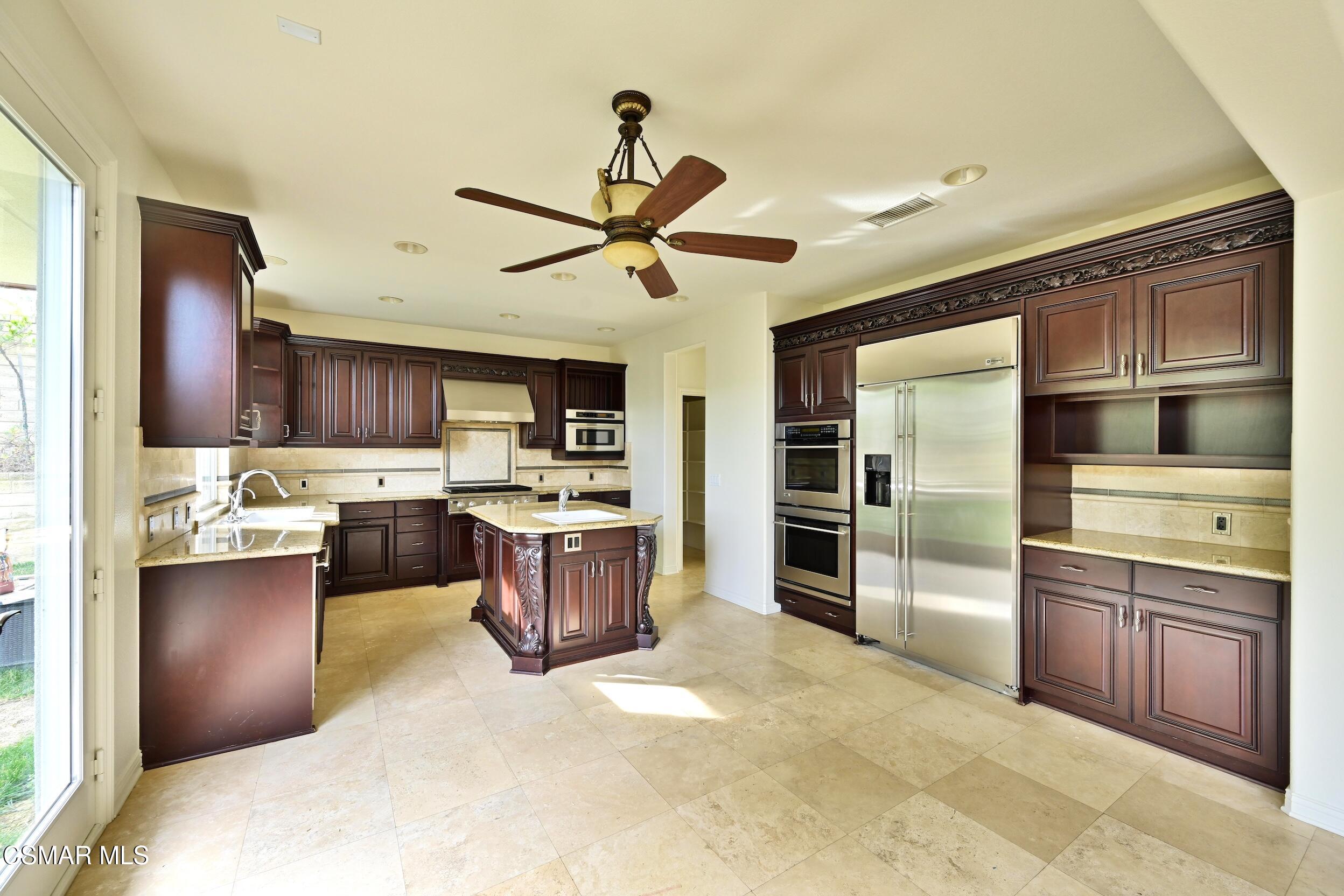 Kitchen with dark wood cabinets, ornate island, stainless steel appliances, and light stone tile floors.