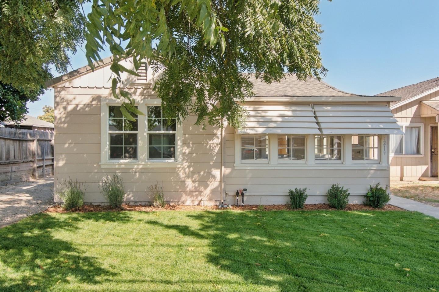 Beige single-story house with a long row of windows beneath a metal awning, shaded by a leafy tree.