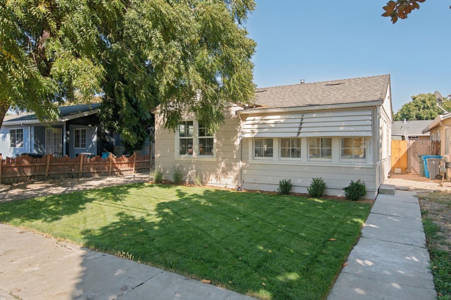 Single-story beige house with wide windows, a green lawn, and a large shade tree.