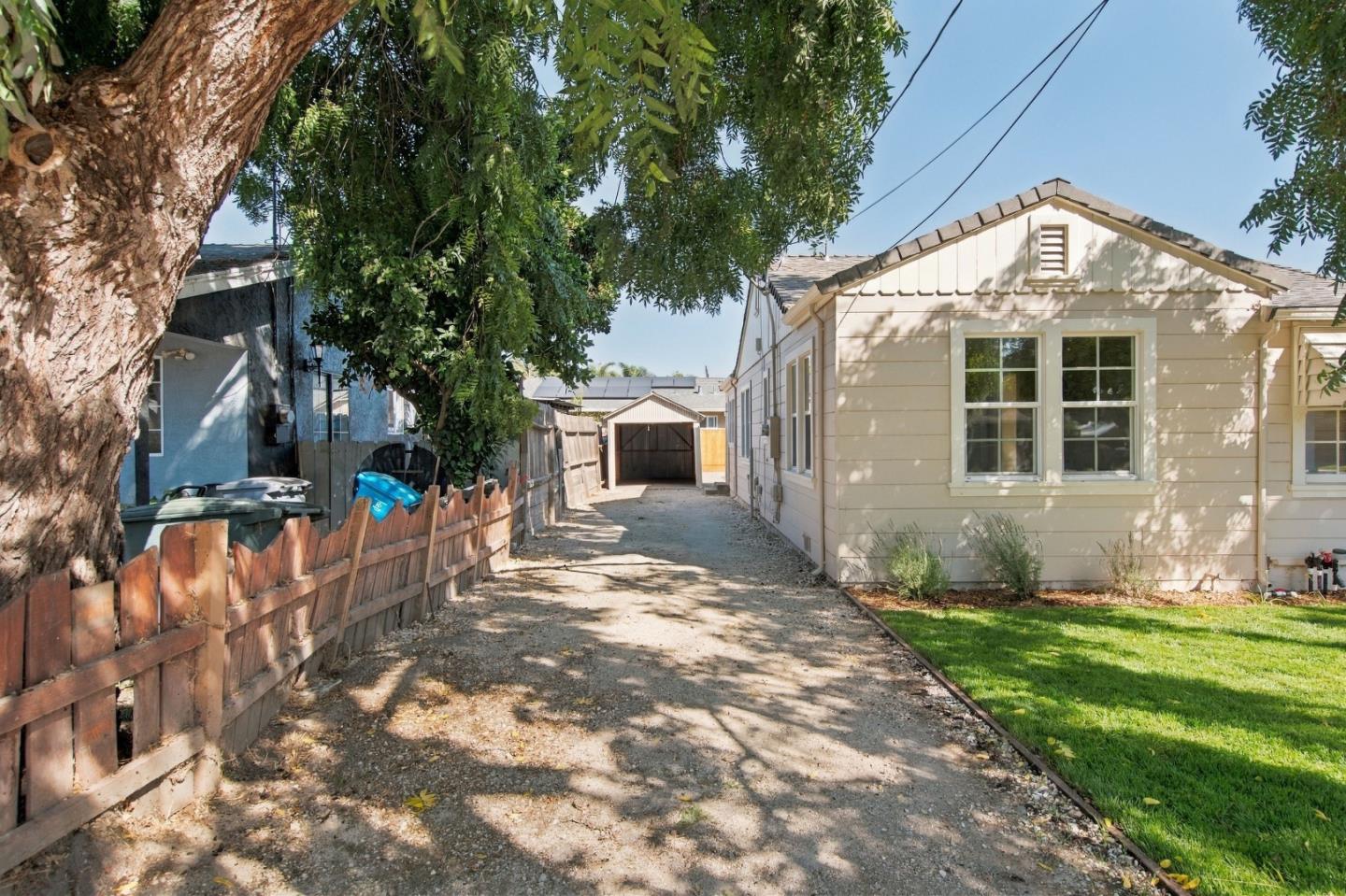 Single-story house beside a gravel driveway leading to a detached shed, shaded by a large tree.