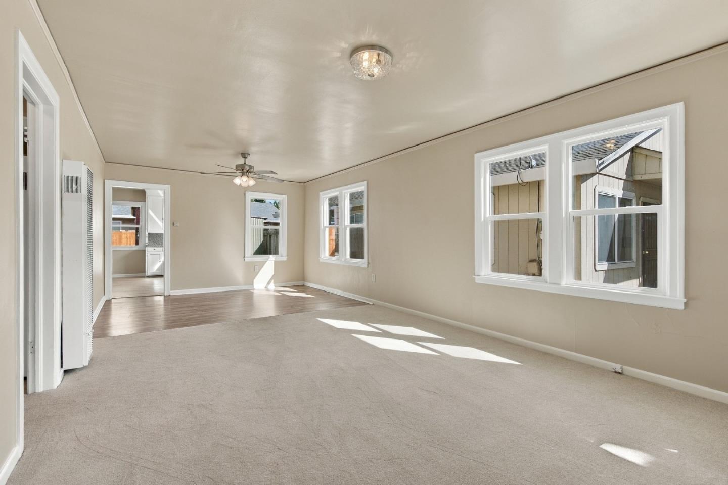 Empty living room with beige carpet, several windows, and a doorway to the kitchen.