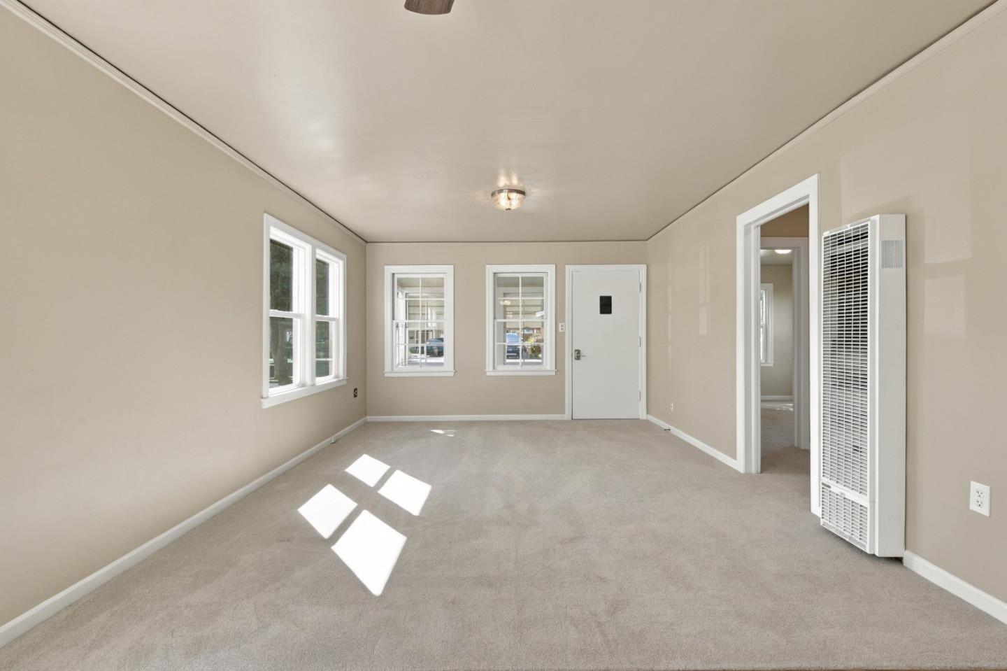 Empty carpeted room with beige walls, multiple windows, a white door, and a wall heater.