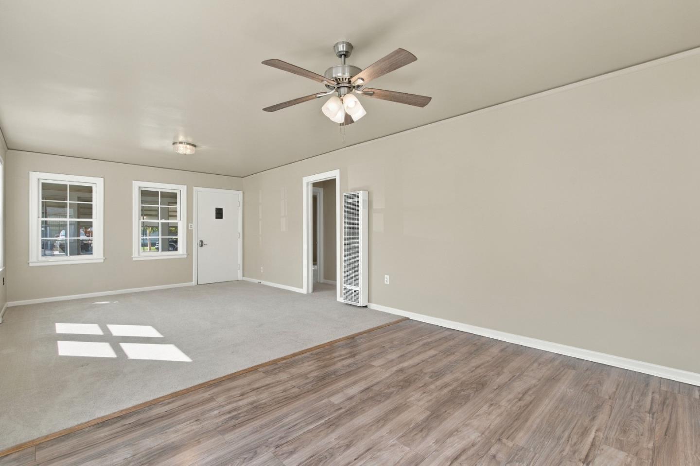Empty living room with gray carpet, wood-look flooring, two windows, and a ceiling fan.