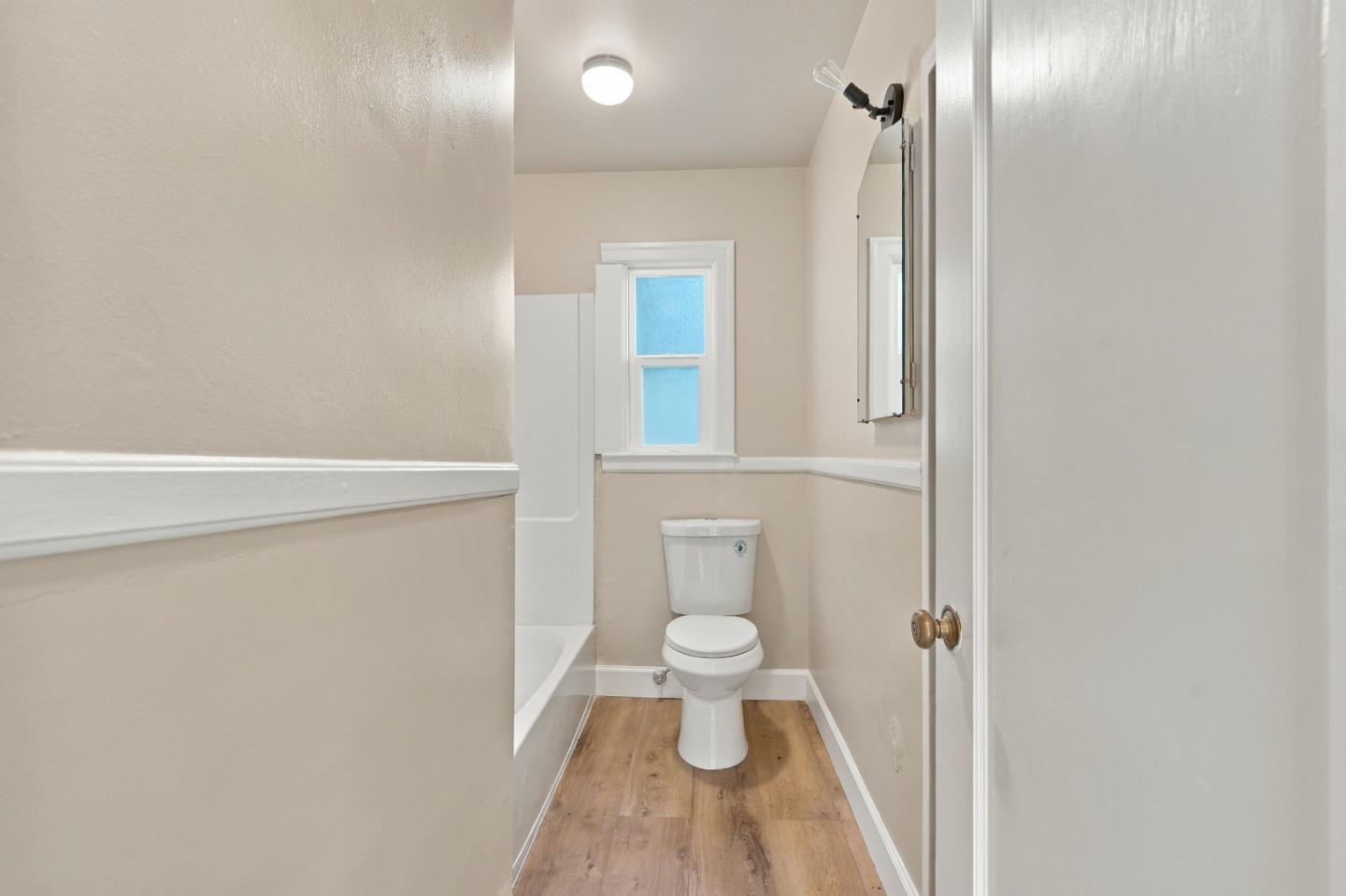 Narrow bathroom with a toilet beneath a small window and wood-look flooring.