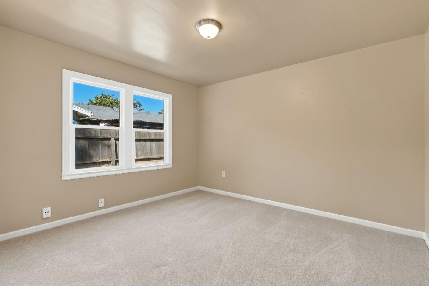 Empty bedroom with beige walls, light carpet, and two windows overlooking a wooden fence.