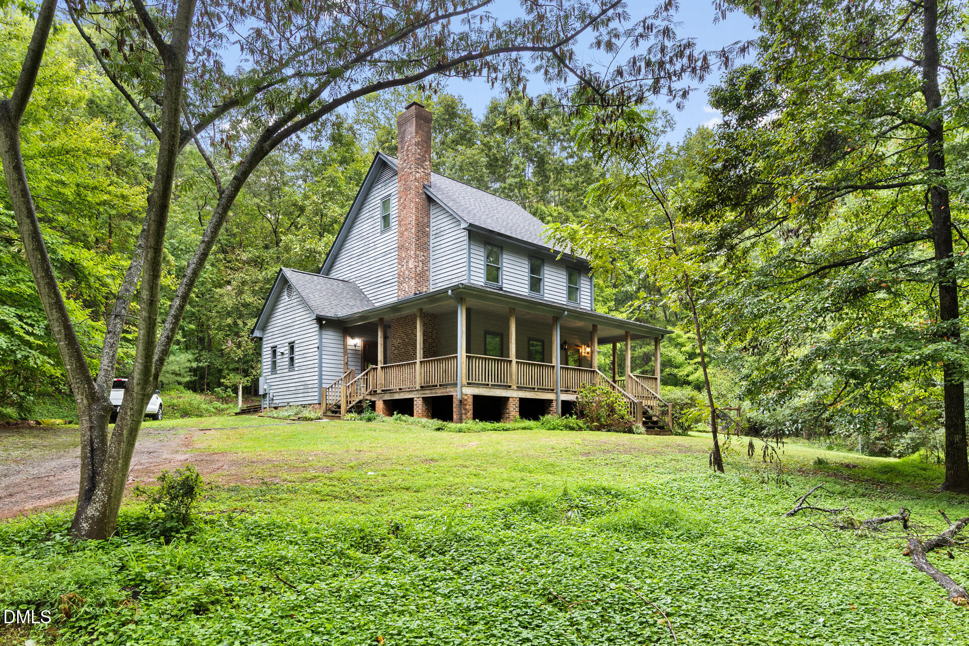 Two-story gray house with a brick chimney and wraparound porch among mature trees.