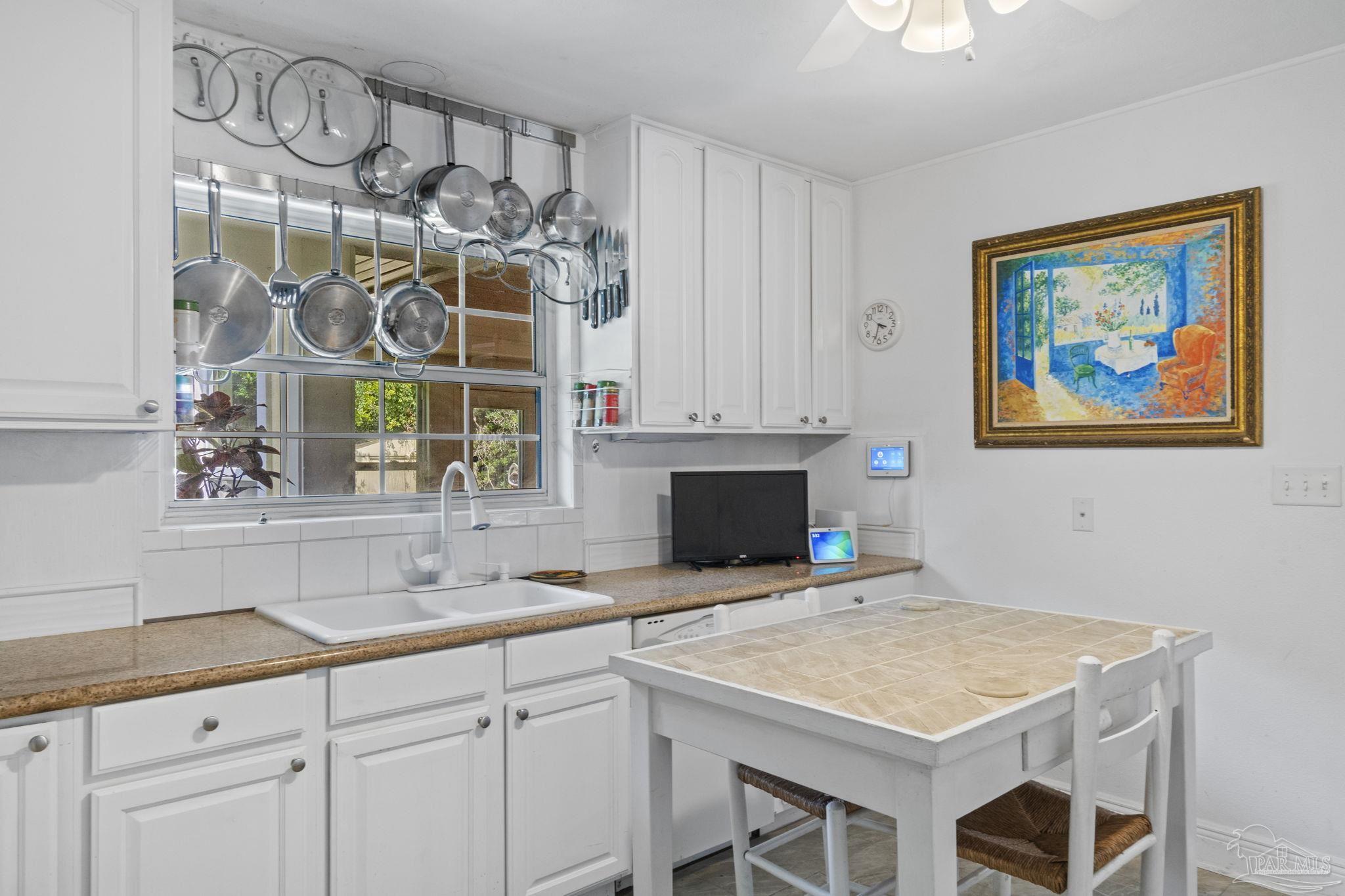 Kitchen with white cabinetry, hanging stainless-steel cookware, a large window, and a tiled table.