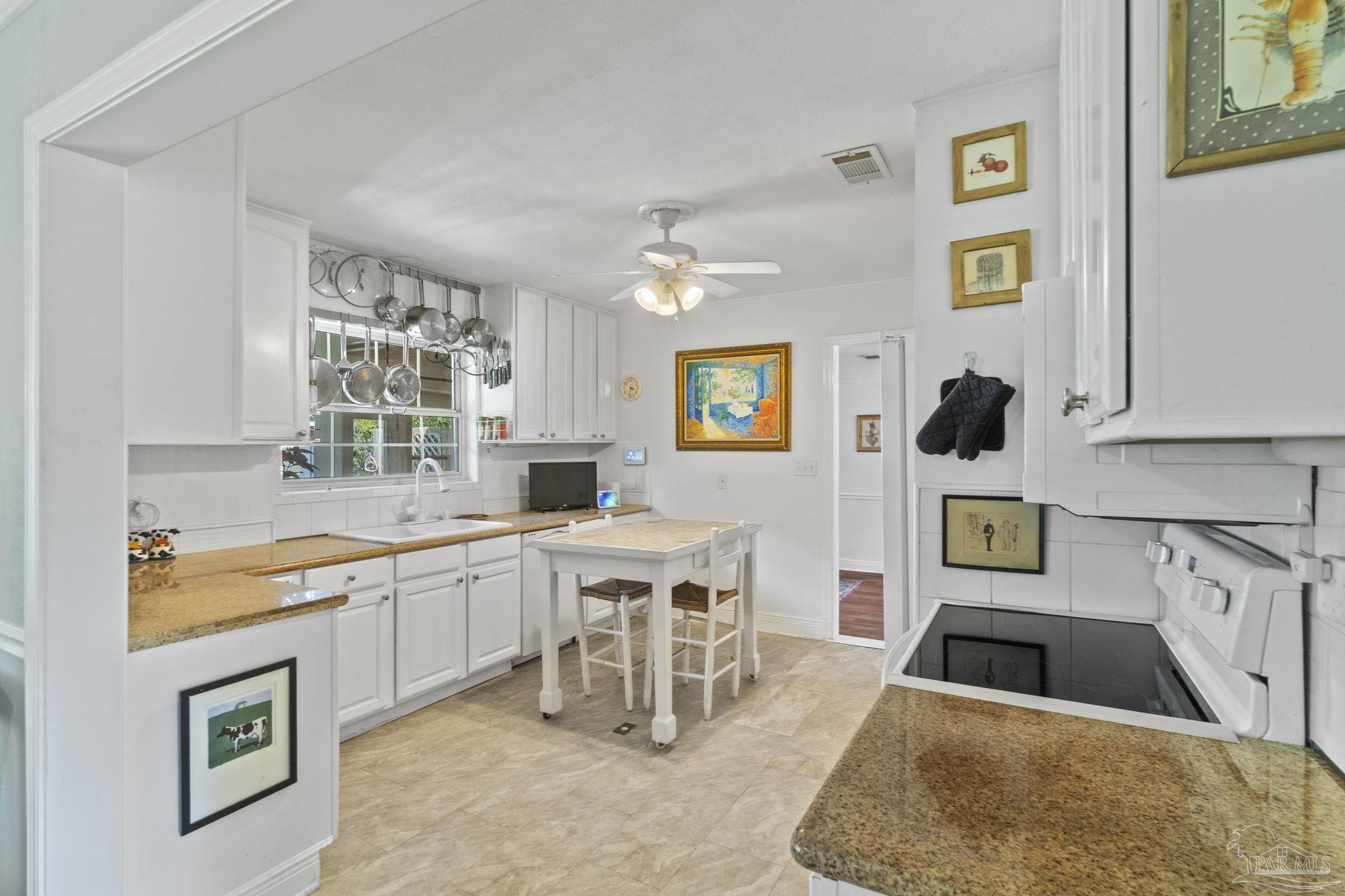 Bright white kitchen with granite counters, hanging pots and pans, and a small table with stools.