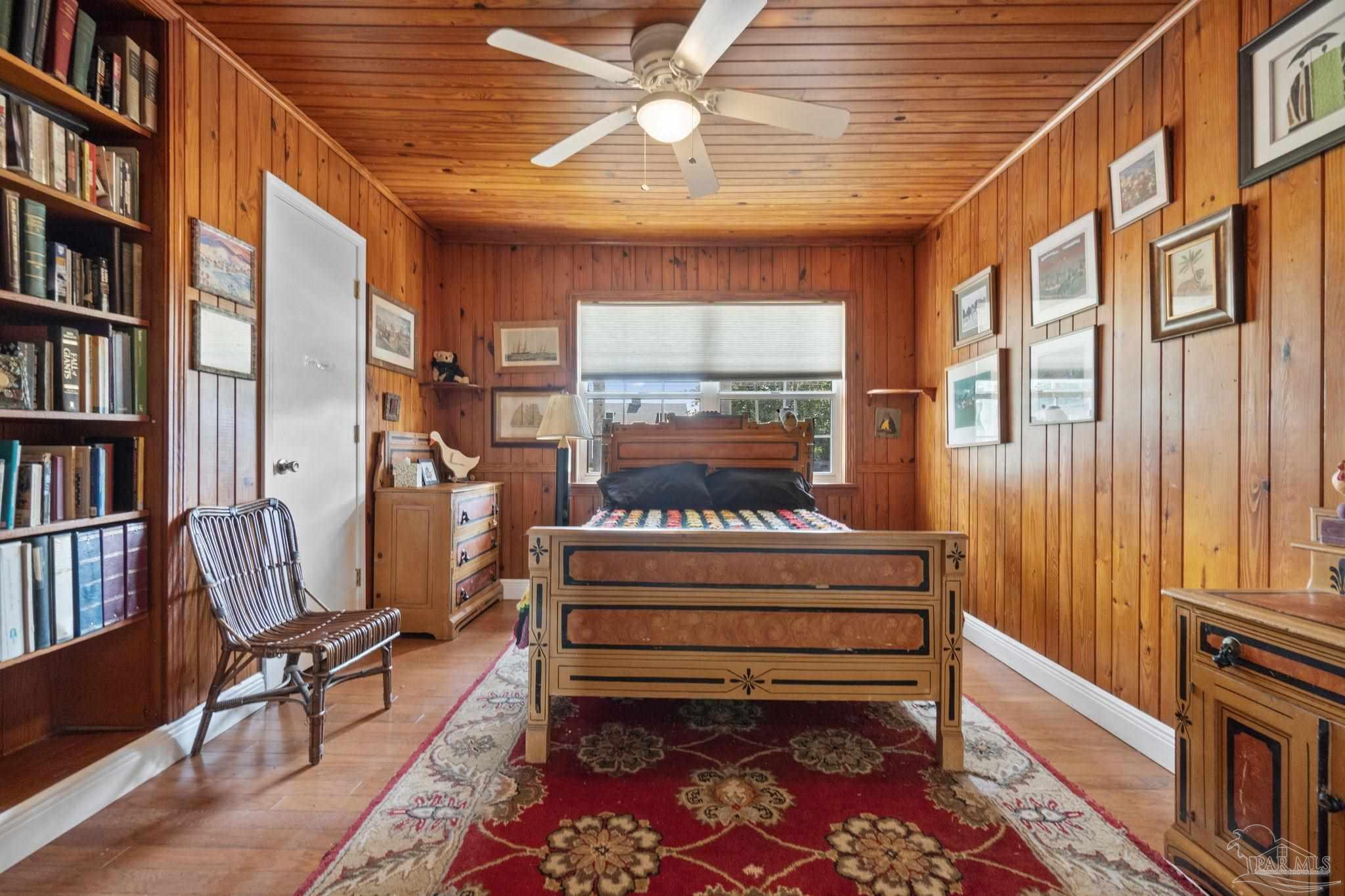Wood-paneled bedroom with a carved bed, built-in bookshelves, framed art, and a red patterned rug.
