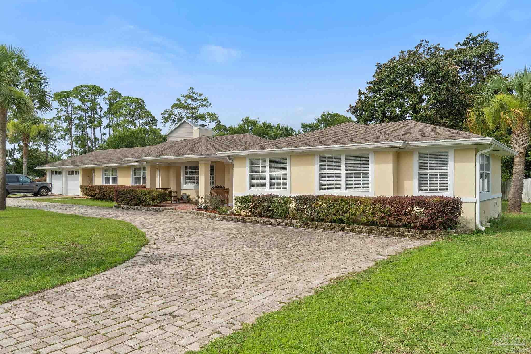 Single-story beige home with a broad curved paver driveway and palm trees.
