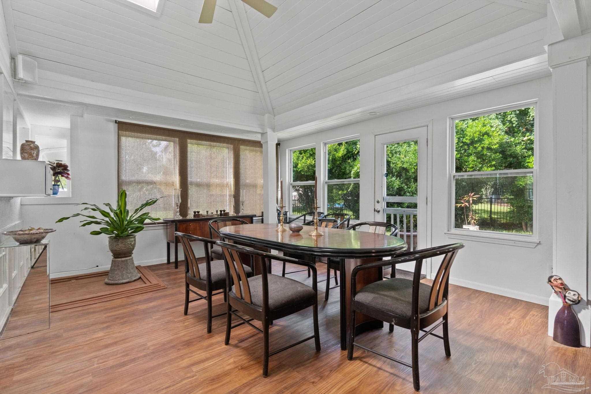 Dining room with an oval table, vaulted ceiling, and windows overlooking leafy greenery.