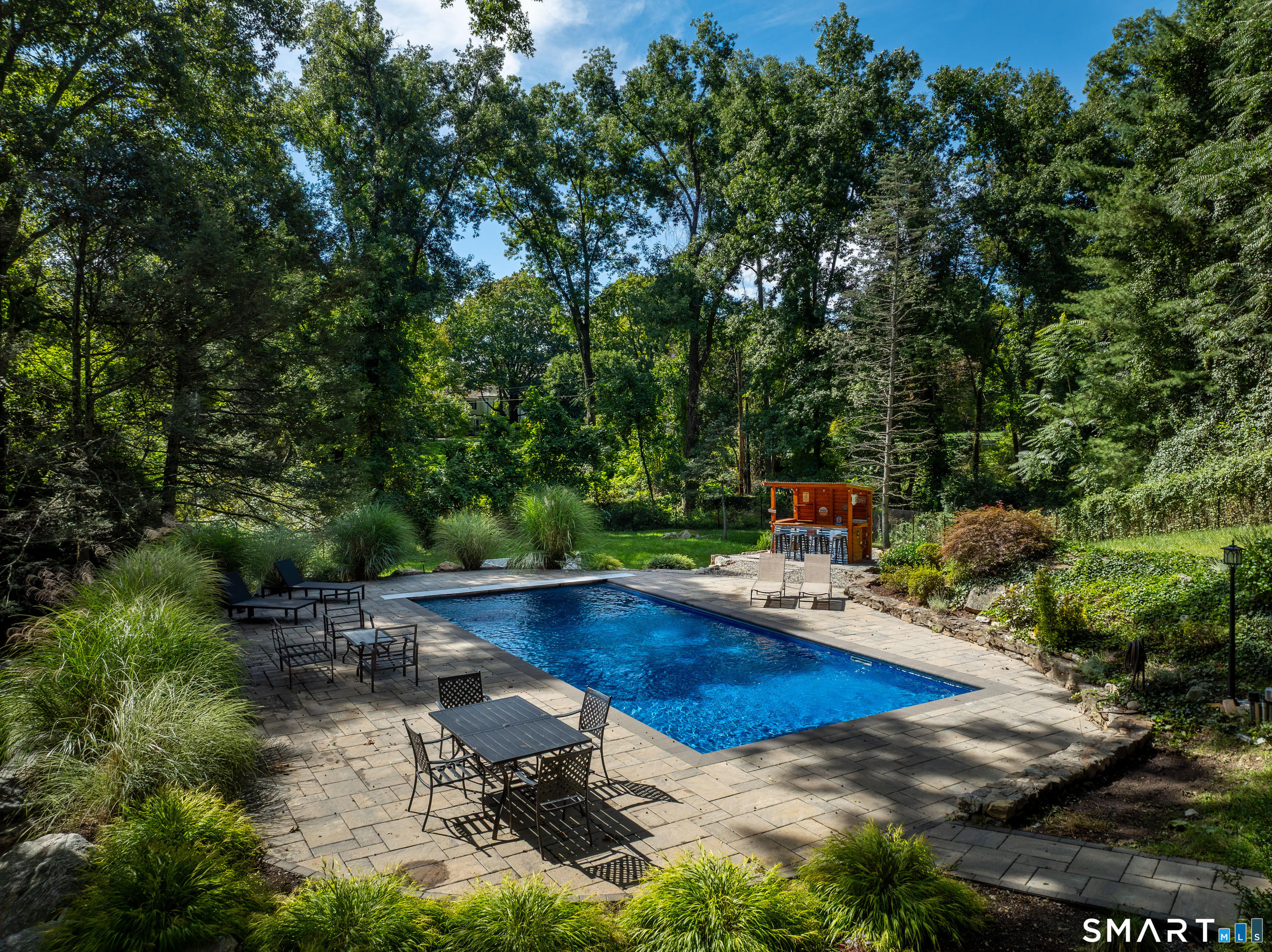 Swimming pool on a stone patio surrounded by mature trees and outdoor seating.