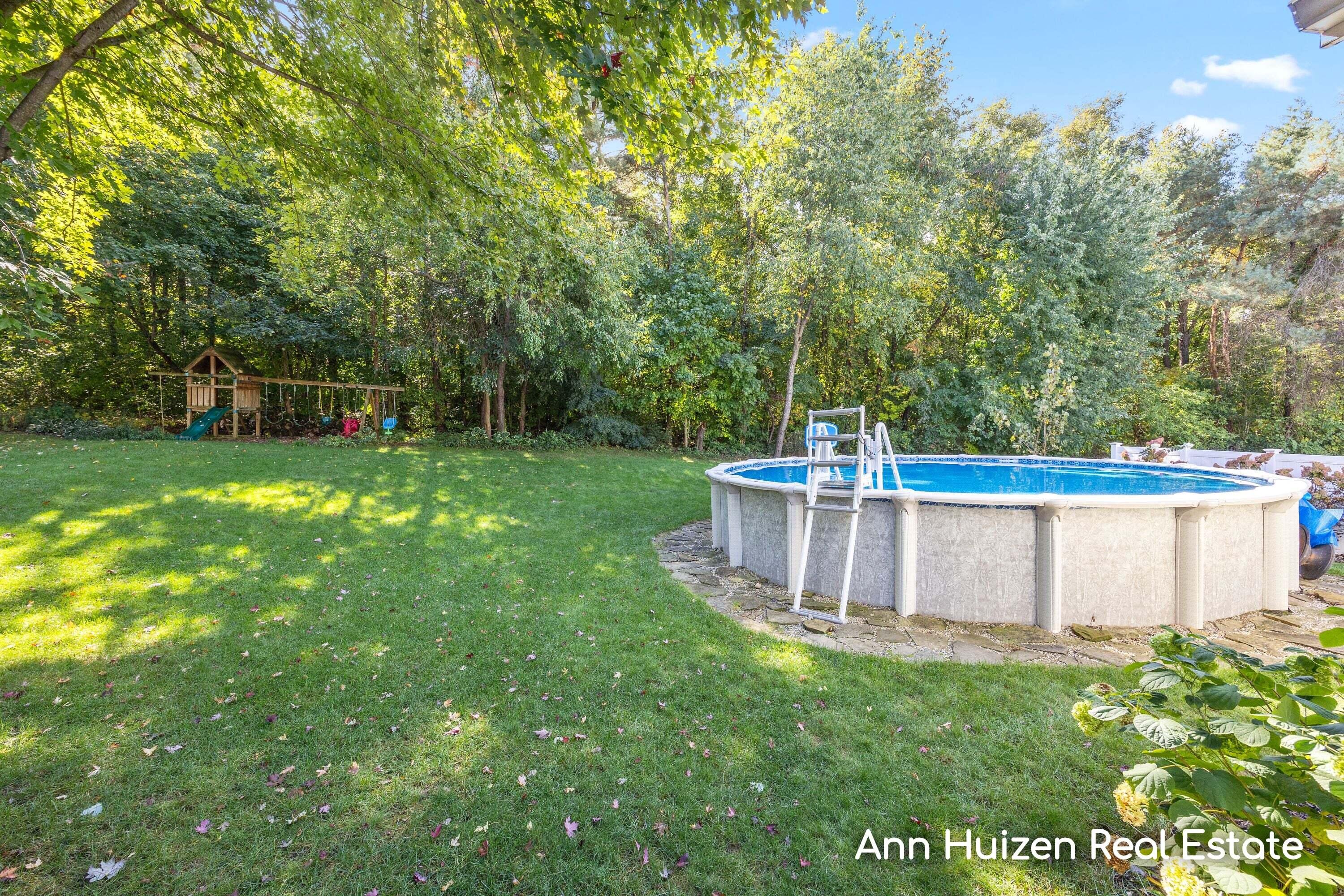Backyard pool beside a broad lawn, with a wooded backdrop and children’s playset.