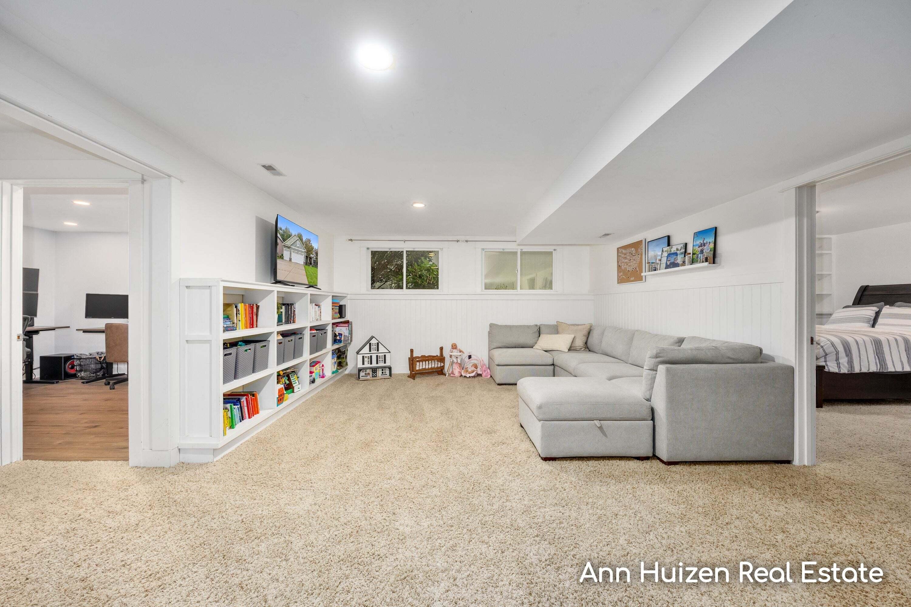Furnished basement family room with a gray sectional, beige carpet, and built-in shelves of books and toys.