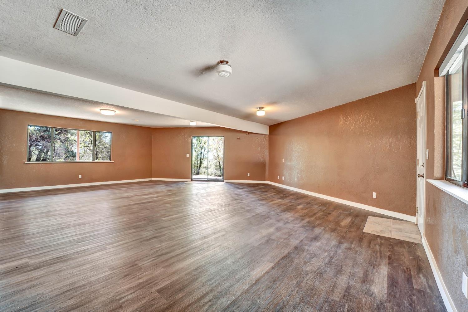 Vacant room with brown walls, plank flooring, broad windows, and a glass door to the trees.