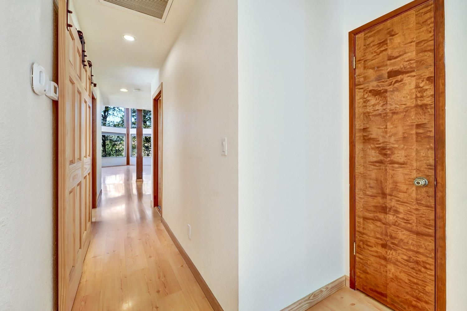 Hallway with light wood floors, paneled closets, a textured wood door, and tall windows overlooking trees.