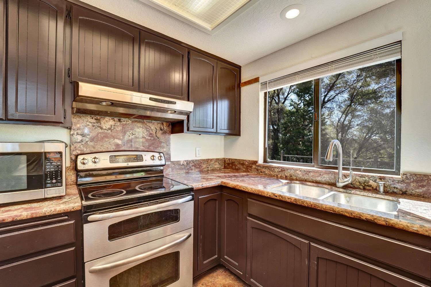 Kitchen with dark wood cabinets, stone counters, stainless steel appliances, and a sink beneath a tree-lined window.