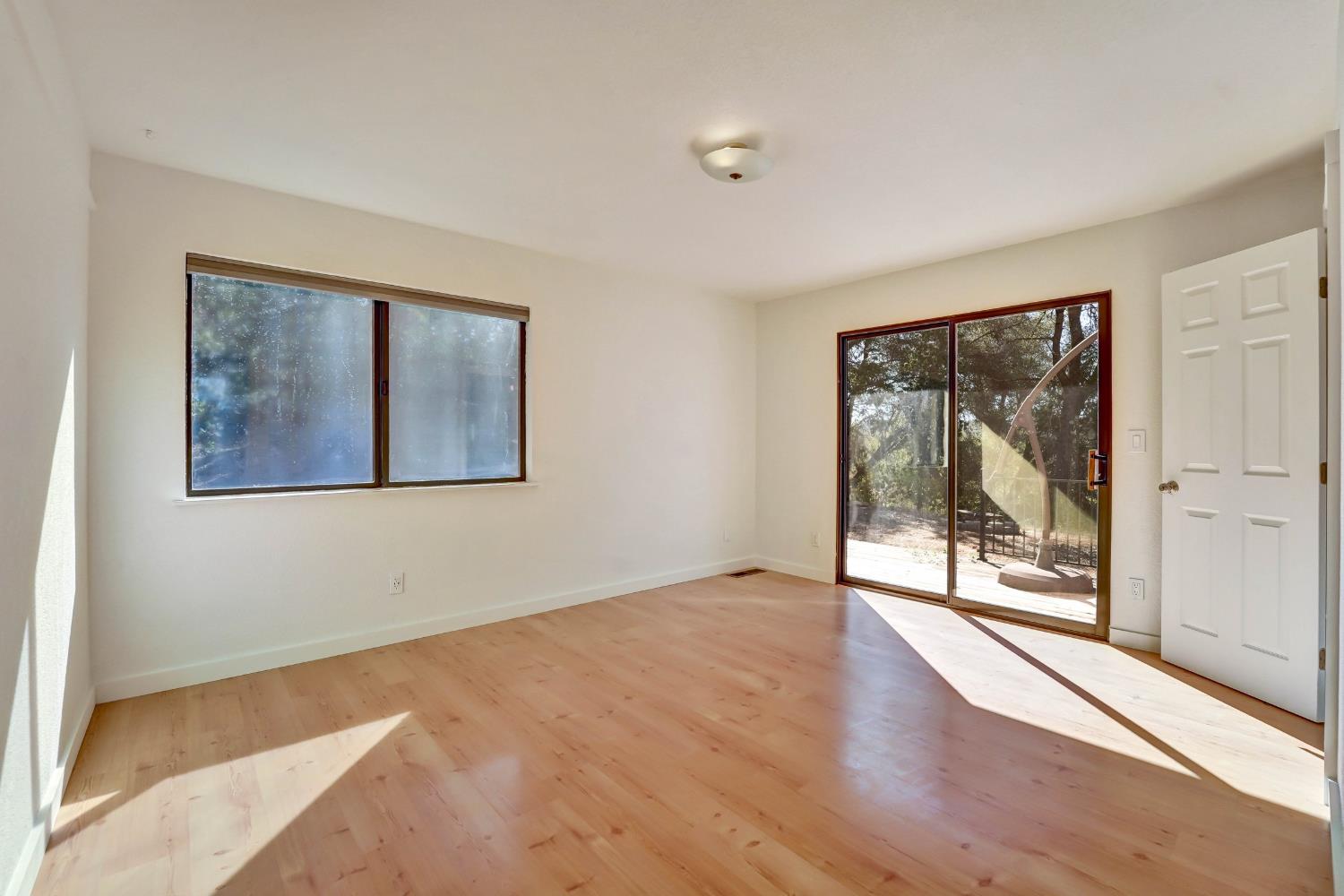 Empty room with light wood floors, a wide window, and a sliding door to a tree-lined deck.