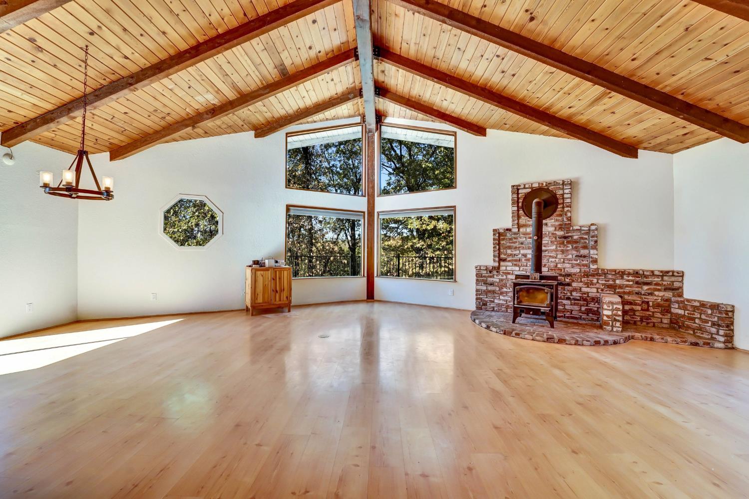 Living room with vaulted wood ceiling, tall tree-facing windows, and a brick fireplace with wood stove.
